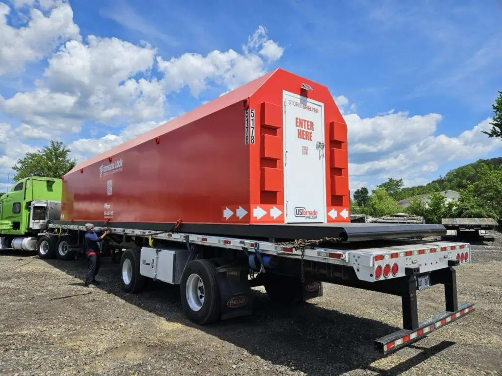 A US Tornado Shelter unit securely loaded on a flatbed trailer, ready for transport. The shelter is bright red with clear signage, engineered for EF-5 tornado protection in commercial or community environments.